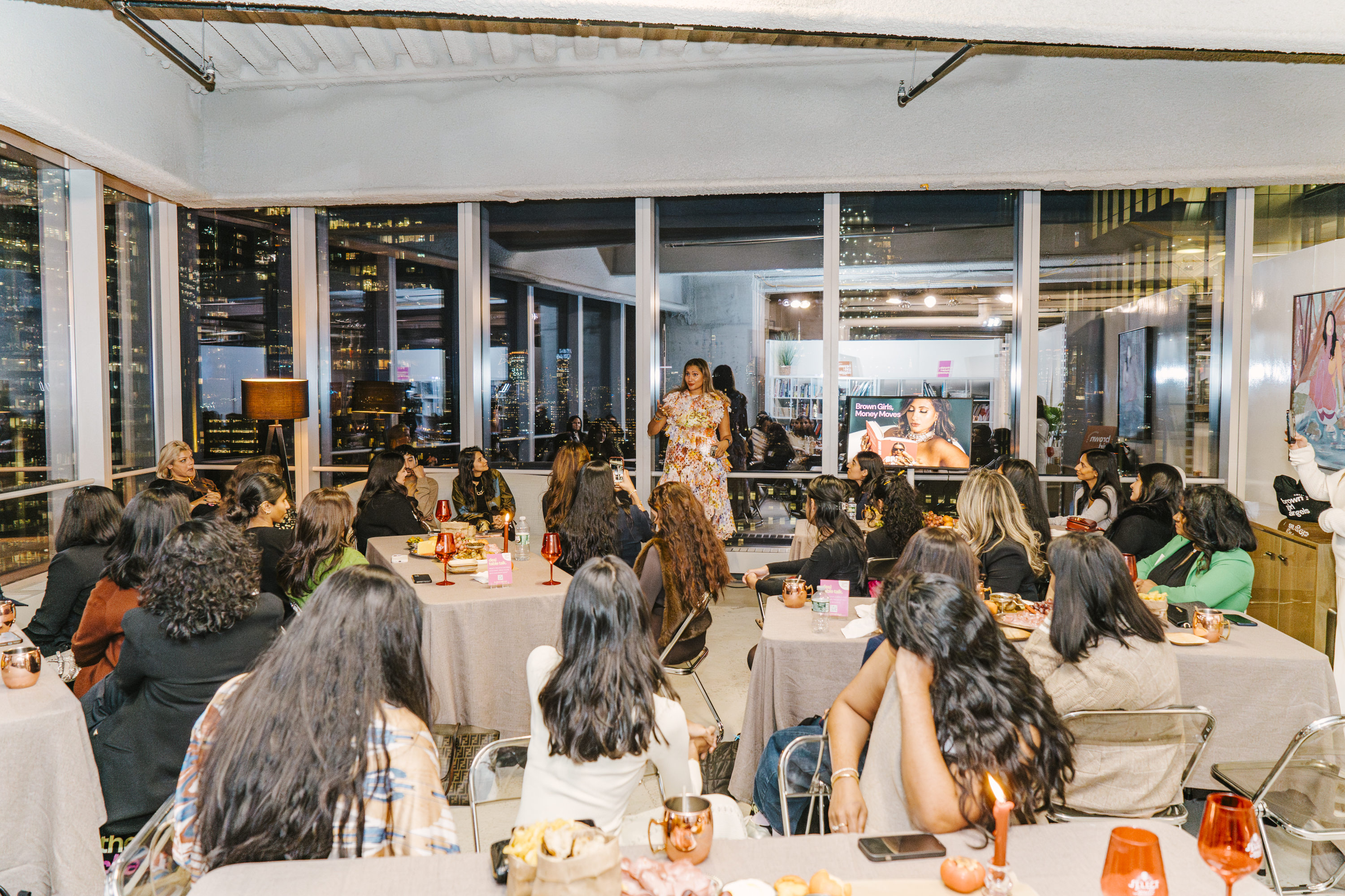 Bhargavi Varma addressing a room of senior South Asian women at a Brown Girl Angels community event in New York &mdash; the Brown Girls, Money Moves brand on screen, full NYC skyline behind her.